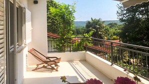 A balcony with a wooden chair, a small table with flowers, and a view of greenery and hills. - Amazing home in Cizici (Cizici)