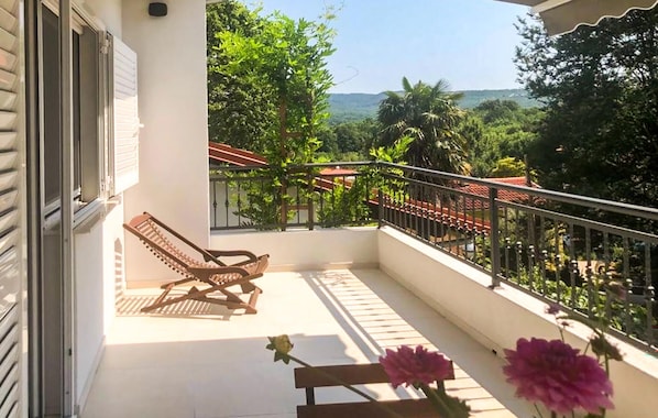 A balcony with a wooden chair, a small table with flowers, and a view of greenery and hills. - Amazing home in Cizici (Cizici)