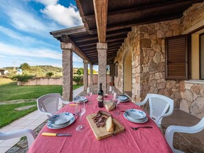 An outdoor dining area with a stone wall, a red tablecloth, white chairs, and a wooden table set with plates, glasses, and a bottle of wine. - Home Thoughtfully Designed for the Comfort (Siniscola)