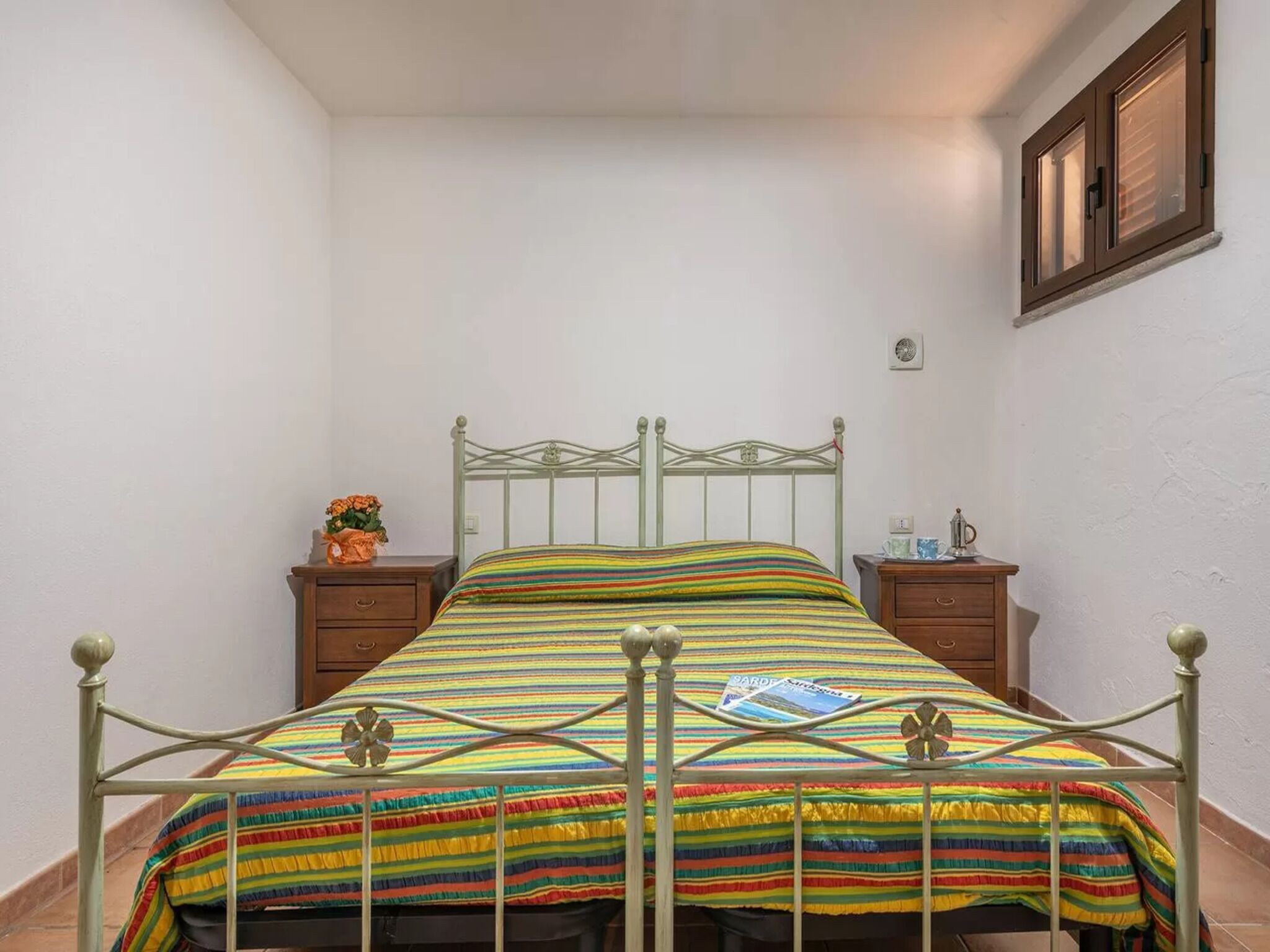 Una cama con estructura metálica, un edredón a rayas de colores y dos mesitas de noche de madera, con un pequeño jarrón de flores en una de ellas.