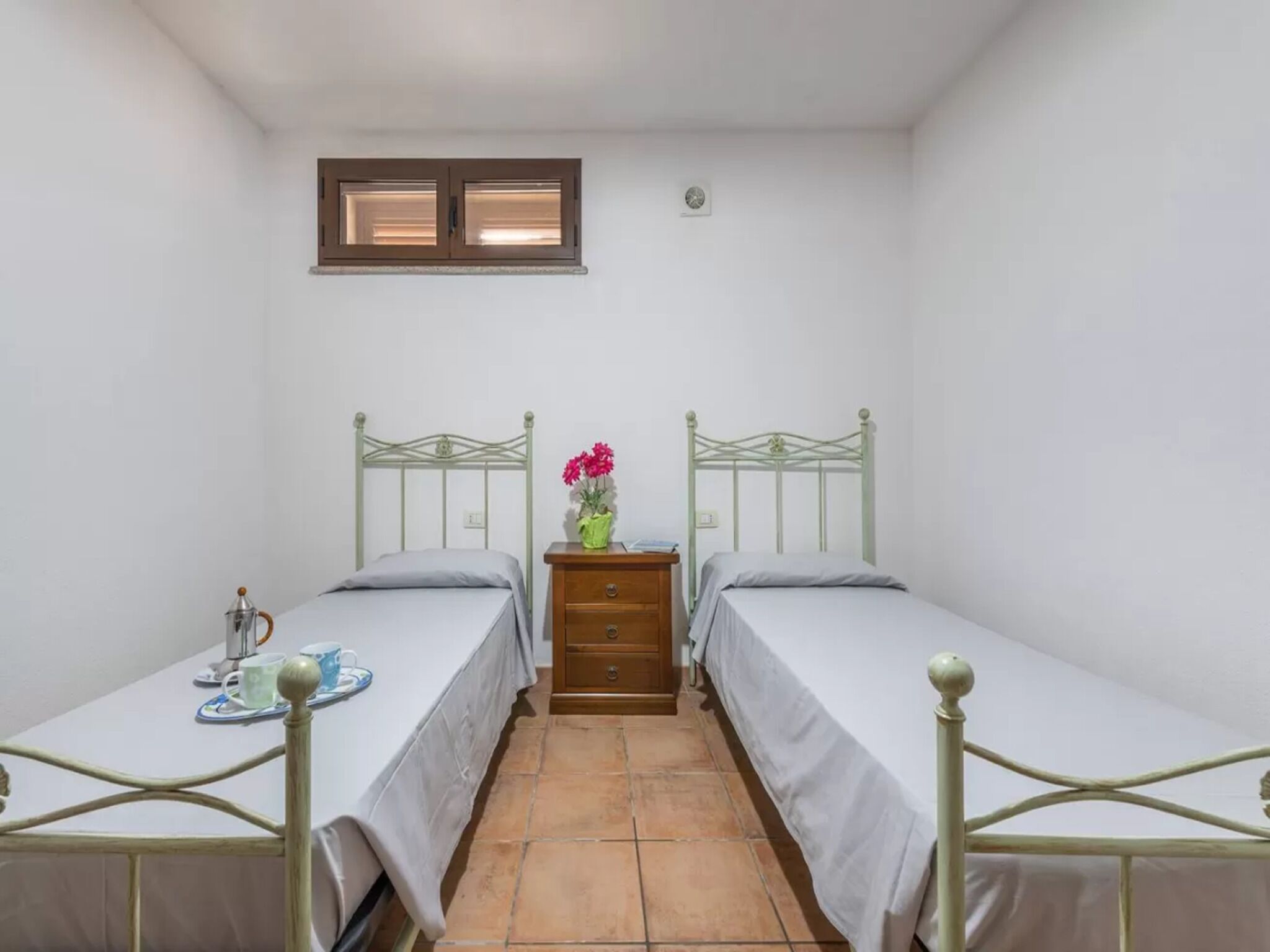 Una habitación pequeña y limpia con dos camas individuales, una mesita de noche con un florero y una ventana sobre las camas.
