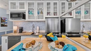 A modern kitchen with white cabinets, a stainless steel refrigerator, and a microwave. There are two dining tables with breakfast items and drinks. - Ocean Oasis - Ocean View At Symphony Beach Club and King Bed (Ormond Beach)