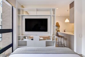 A modern bedroom with a large TV mounted on a white shelving unit, a bed with a grey and white patterned bedspread, a small kitchen area with a bar stool, and a window overlooking a brick building. - w Stylish Radiant Loft in Chapinero (Bogotá)