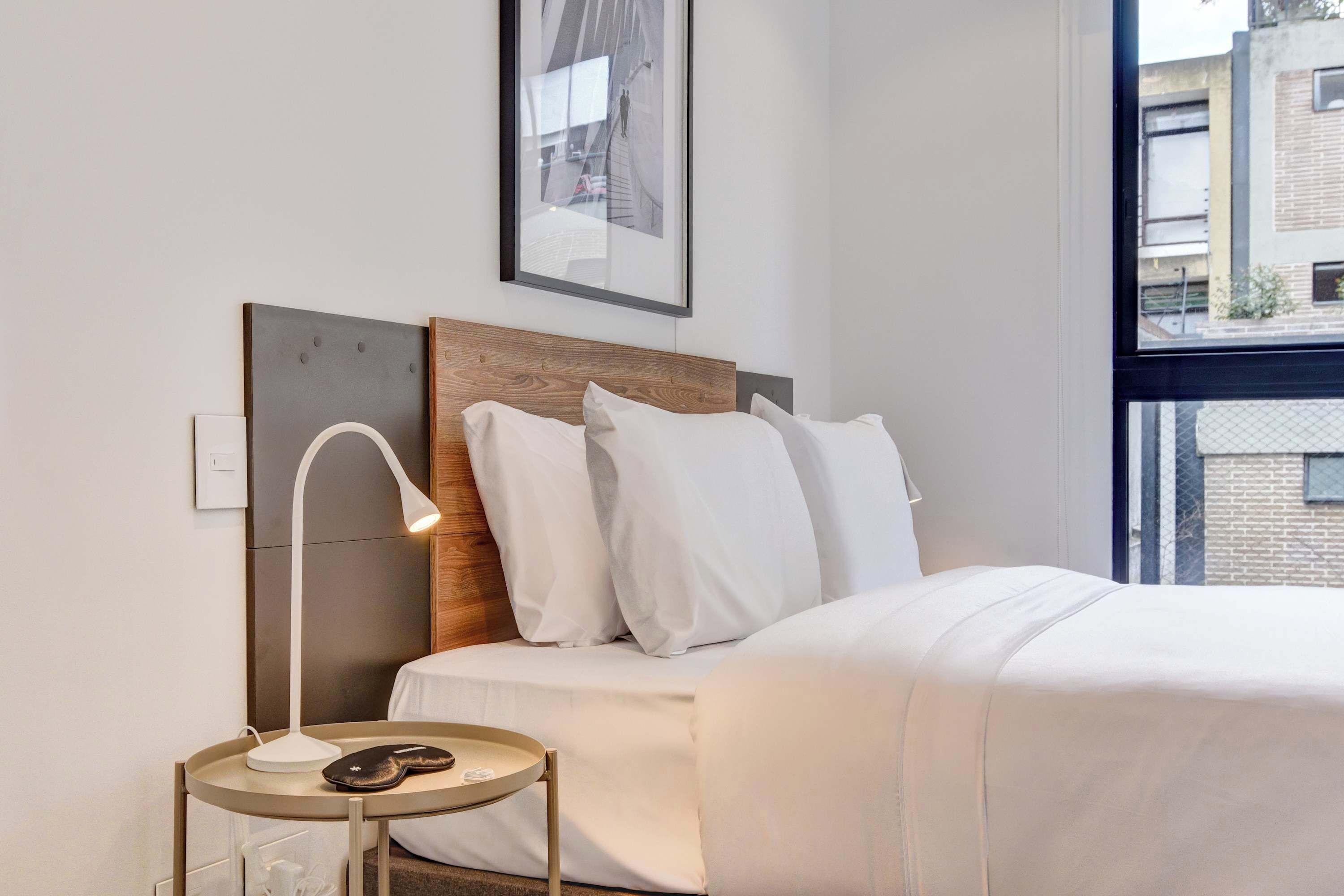 Ein modernes Hotelzimmer mit einem ordentlich bezogenen Bett, einem Nachttisch mit Lampe und einem gerahmten Bild an der Wand.
