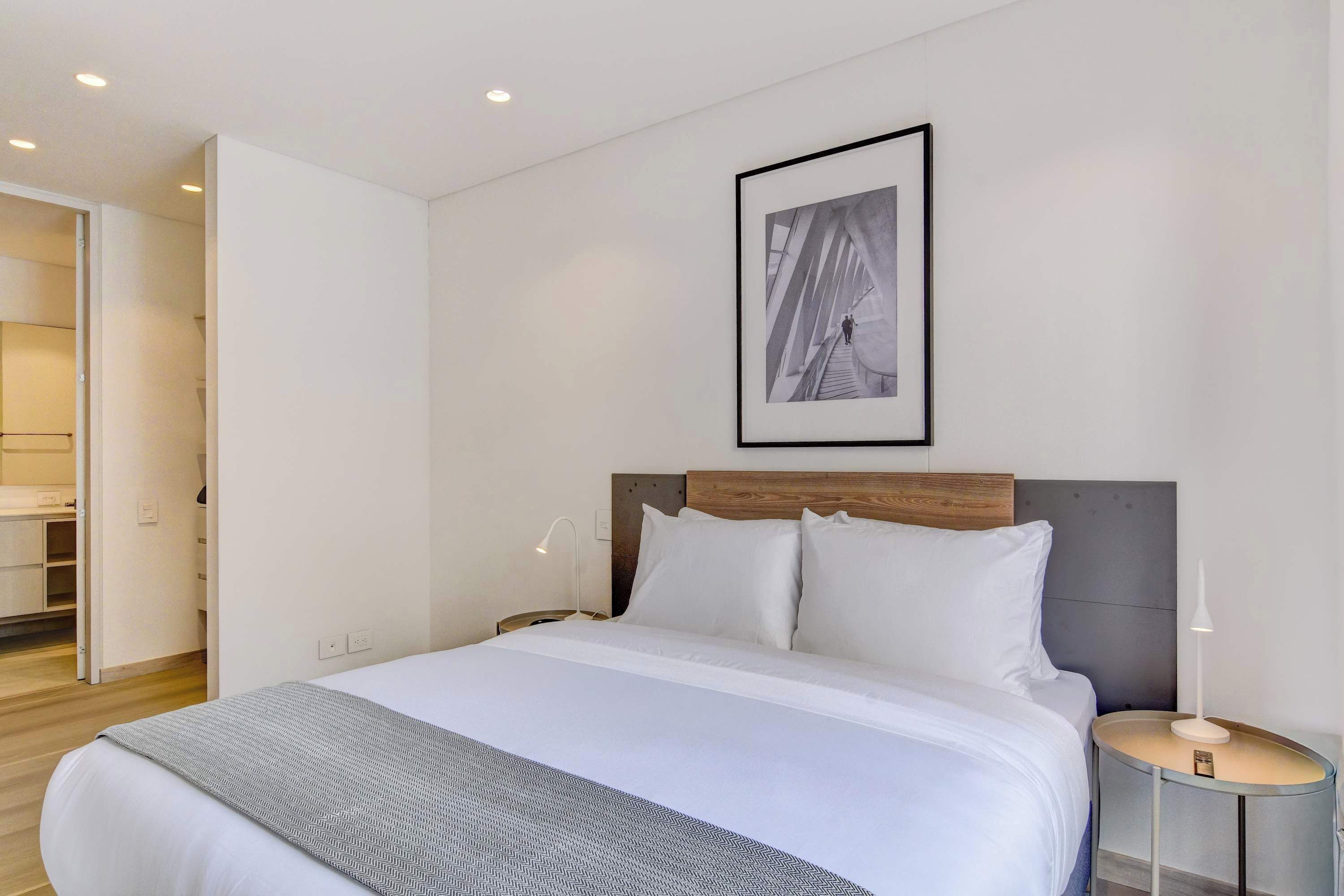 A modern hotel room with a neatly made bed, a bedside table with a lamp, and a framed picture on the wall.