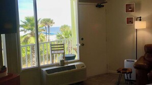 Ein Hotelzimmer mit Bett, Fernseher, Fenster mit Blick auf Palmen und Meer, Sessel, kleiner Tisch und Lampe.