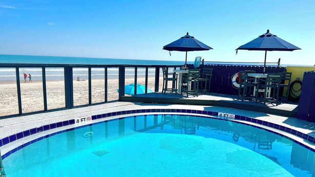 Ein Swimmingpool mit einer 1,2-Meter-Wassertiefenanzeige, Blick auf den Strand und Liegemöglichkeiten mit Sonnenschirmen.
