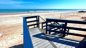 Ein hölzerner Steg führt zu einem Strand mit blauem Himmel und ruhigem Wasser.