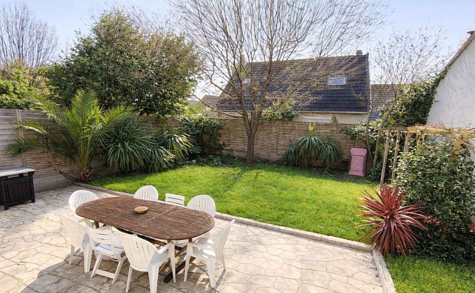 Une terrasse avec une table ronde et des chaises, entourée d’une cour verdoyante et de différentes plantes.