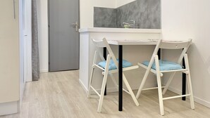 Eine kleine Küche mit einer weißen Arbeitsplatte, zwei weißen Stühlen mit blauen Kissen und einer grauen Tür.