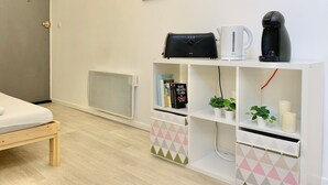 Ein modernes Hotelzimmer mit einem Bett, einem weißen Regal mit Kaffeemaschine, Wasserkocher und einer kleinen Pflanze. An der Wand hängen zwei gerahmte Bilder und darunter ein Heizkörper.