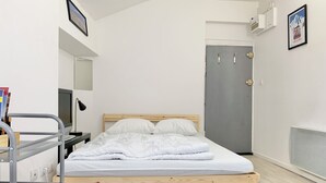 Ein Einzelbett mit Holzrahmen und weißer Bettwäsche, ein Schreibtisch mit Computer und Büchern sowie eine graue Tür.