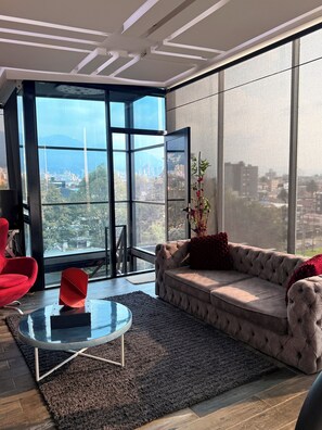 Una sala de estar moderna con un ventanal grande, un sofá capitonado, un sillón rojo y una mesa de centro de vidrio.