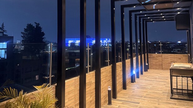 A rooftop terrace with a wooden floor, metal railings, and a view of city buildings at night. - MOVISTAR ARENA PENTHOUSE (Bogotá, D.C.)