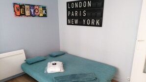 Ein kleines, ordentlich bezogenes Bett mit einer türkisfarbenen Tagesdecke und zwei Kissen, eines davon weiß mit einem schwarzen Herz. An der Wand hängt ein Schild mit der Aufschrift 'LONDON PARIS NEW YORK'.
