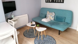 Ein modernes Wohnzimmer mit einem petrolfarbenen Sofa, einem weißen Couchtisch, einem kleinen Beistelltisch und einem an der Wand befestigten Fernseher.