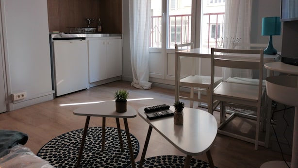 Eine moderne Küche mit weißen Schränken, einer Uhr und einem Fenster mit Vorhängen. Es gibt einen Essbereich mit Tisch und Stühlen, einem kleinen Teppich und einer Pflanze auf dem Tisch.