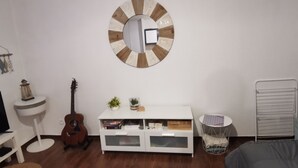 Una habitación con un tocador blanco, una guitarra, un espejo redondo, un armario blanco con libros y plantas, y un radiador.