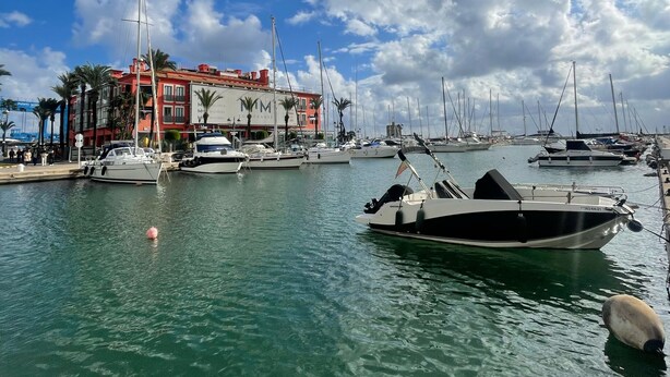A marina with boats docked, a building with red and white facade, and a clear blue sky with clouds.