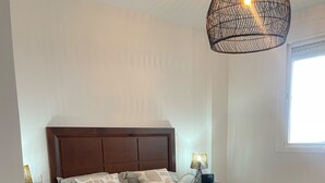 A neatly made bed with a green bedspread and multiple pillows, a wooden nightstand with a lamp, and a hanging light fixture above the bed.