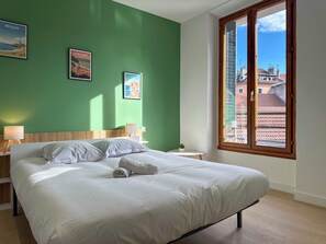 En pænt redt seng med hvide sengetøj og puder, et lille natbord med lampe, og to billeder i rammer på den grønne væg.