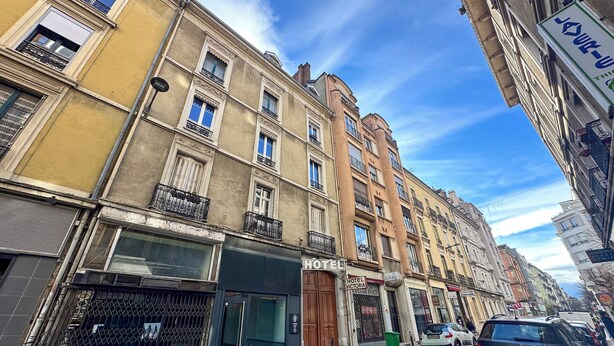 Un immeuble à plusieurs étages, avec une enseigne indiquant « HÔTEL », une entrée bien visible et une vue sur la rue où sont garées des voitures.