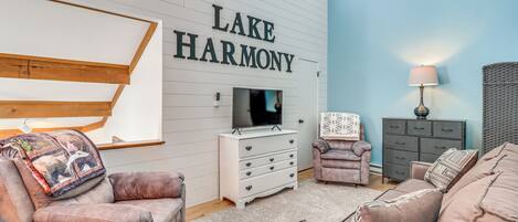 Uma sala de estar com televisão, cômoda branca, sofá marrom e uma parede azul com a inscrição "LAKE HARMONY".