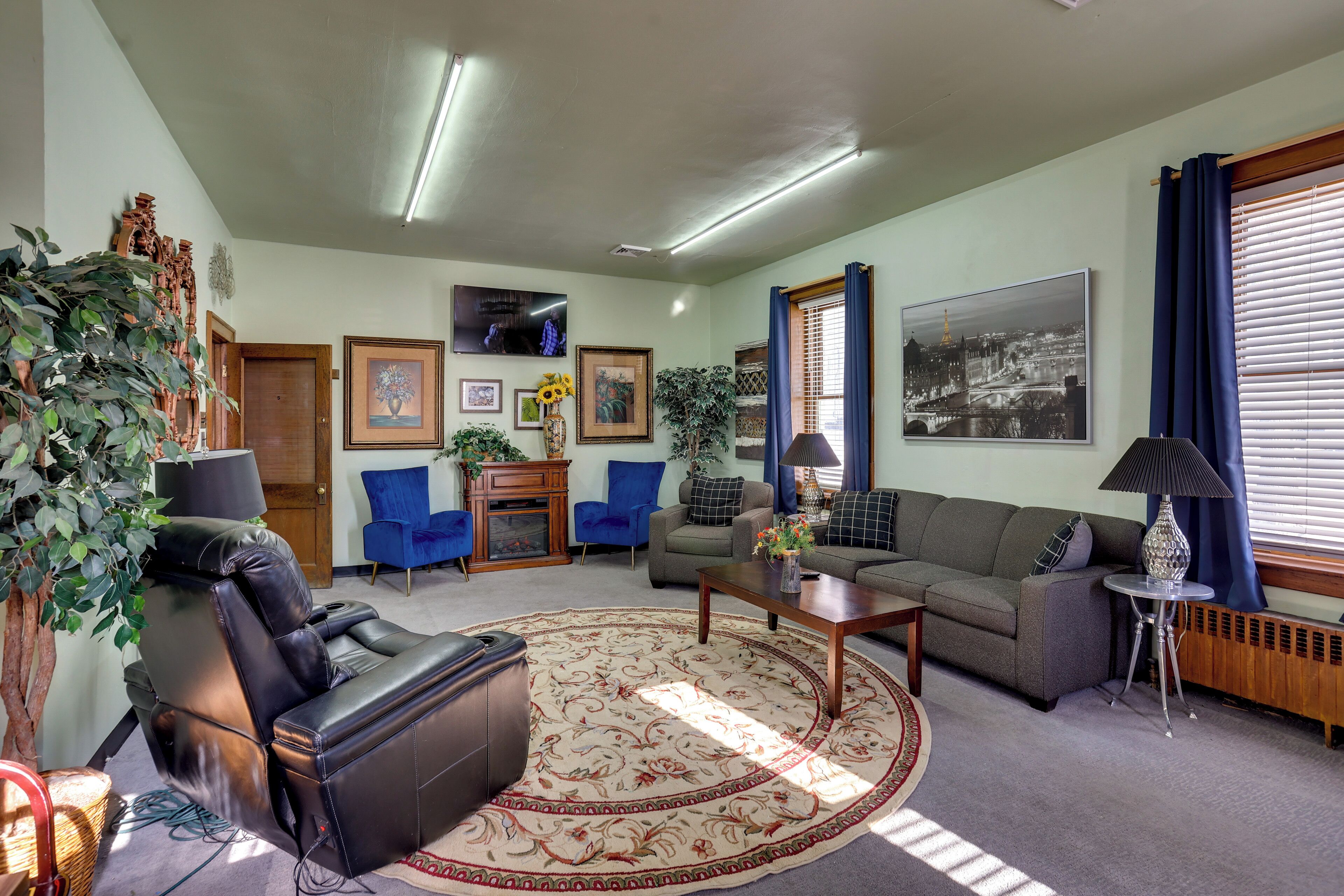 A living room with a leather sofa, a coffee table, and a patterned rug. There are two blue chairs, a fireplace, and a large window with curtains.