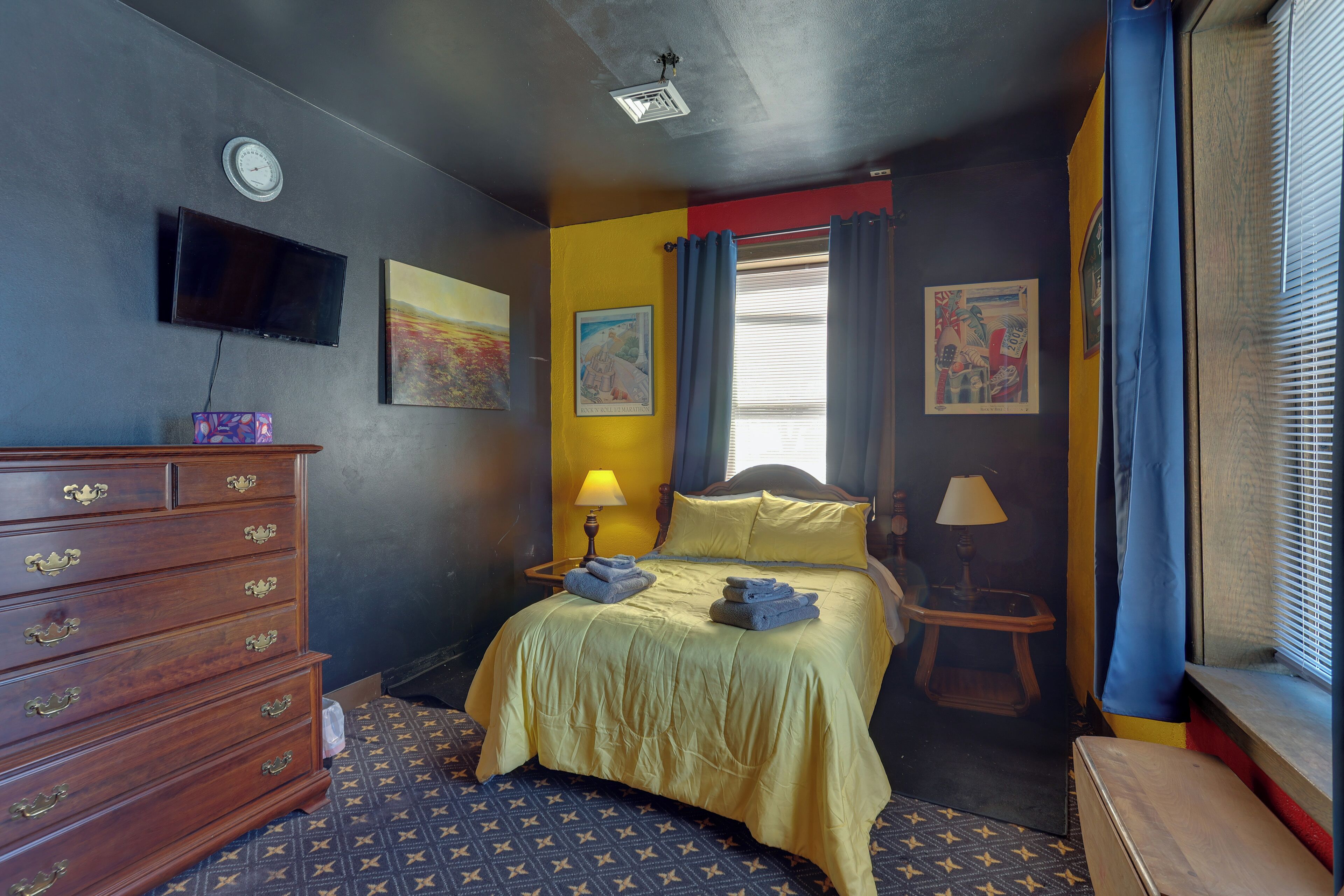 A hotel room with a bed, dresser, wall-mounted TV, artwork, and a window with curtains.