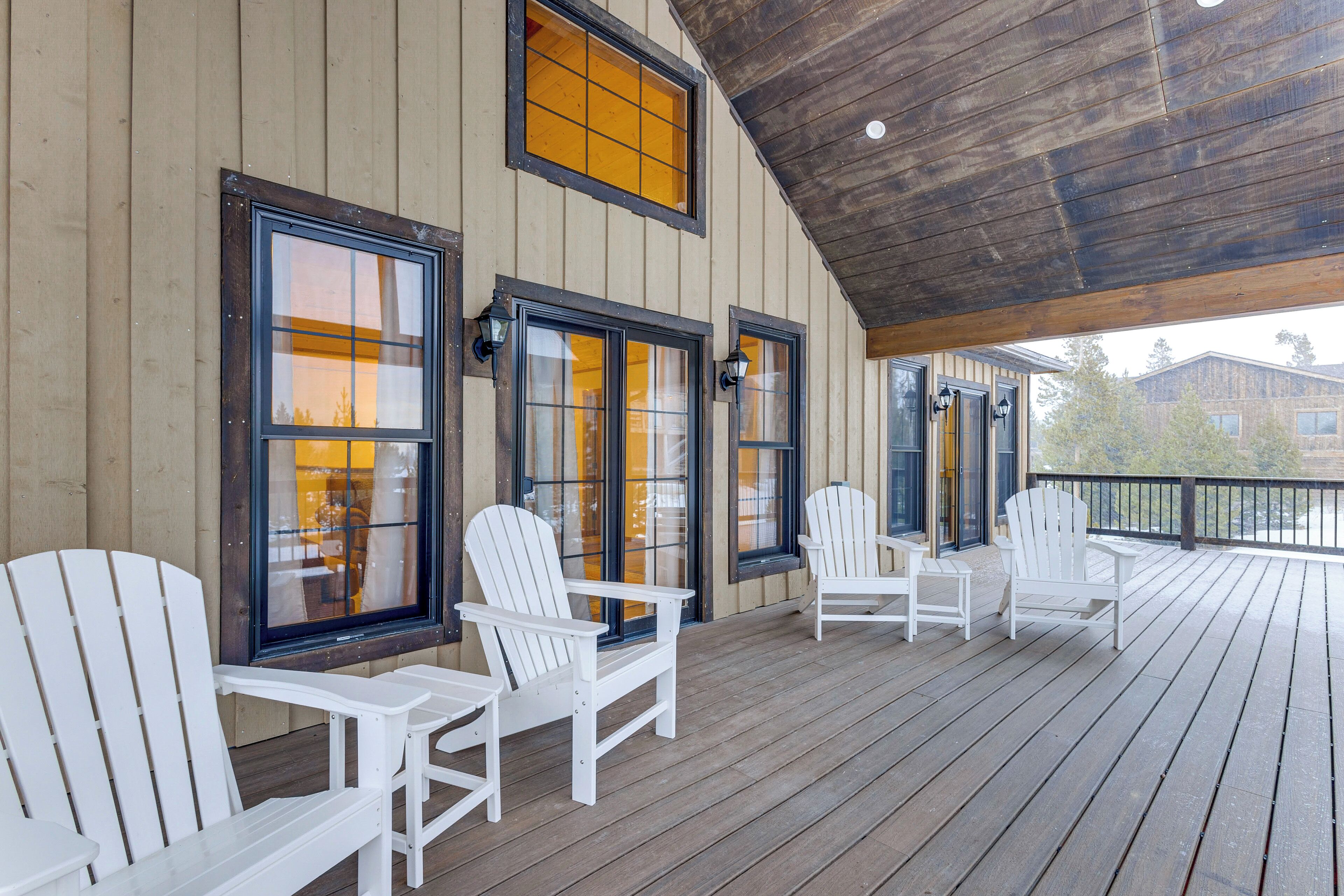 En træterrasse med hvide Adirondack-stole og store vinduer med mørke rammer.