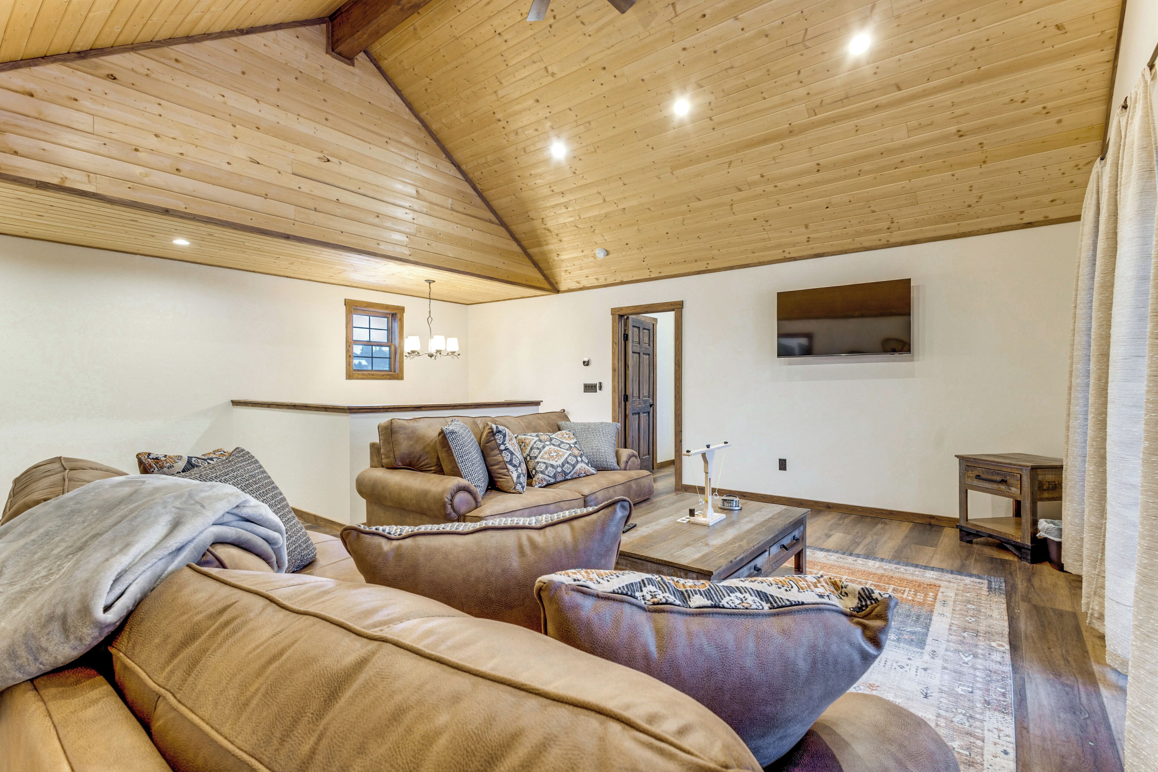 A spacious living room with a high wooden ceiling, leather sofas, a coffee table, and a television mounted on the wall.