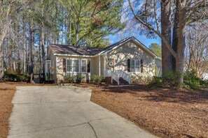 A single-story house with a front porch, a driveway, and a wooded area.
