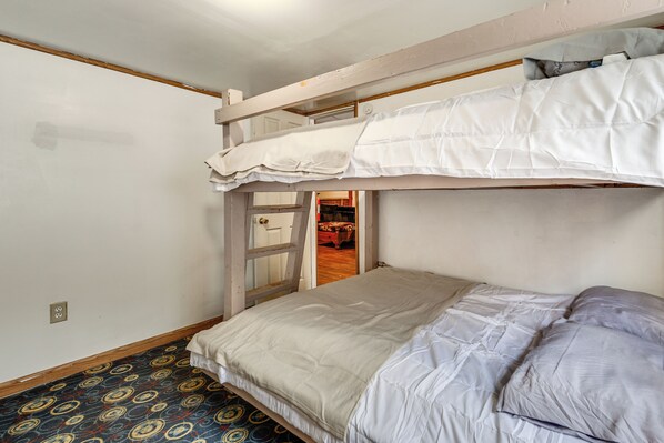 Habitación con literas, alfombra estampada, una puerta que da a otra habitación y una pared blanca con toma de corriente.