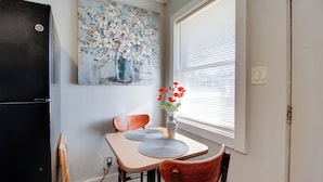 A dining area with a table, chairs, a vase with flowers, a painting on the wall, and a window with blinds. - Cozy Huntsville Studio Near Lowe Mill (Huntsville)