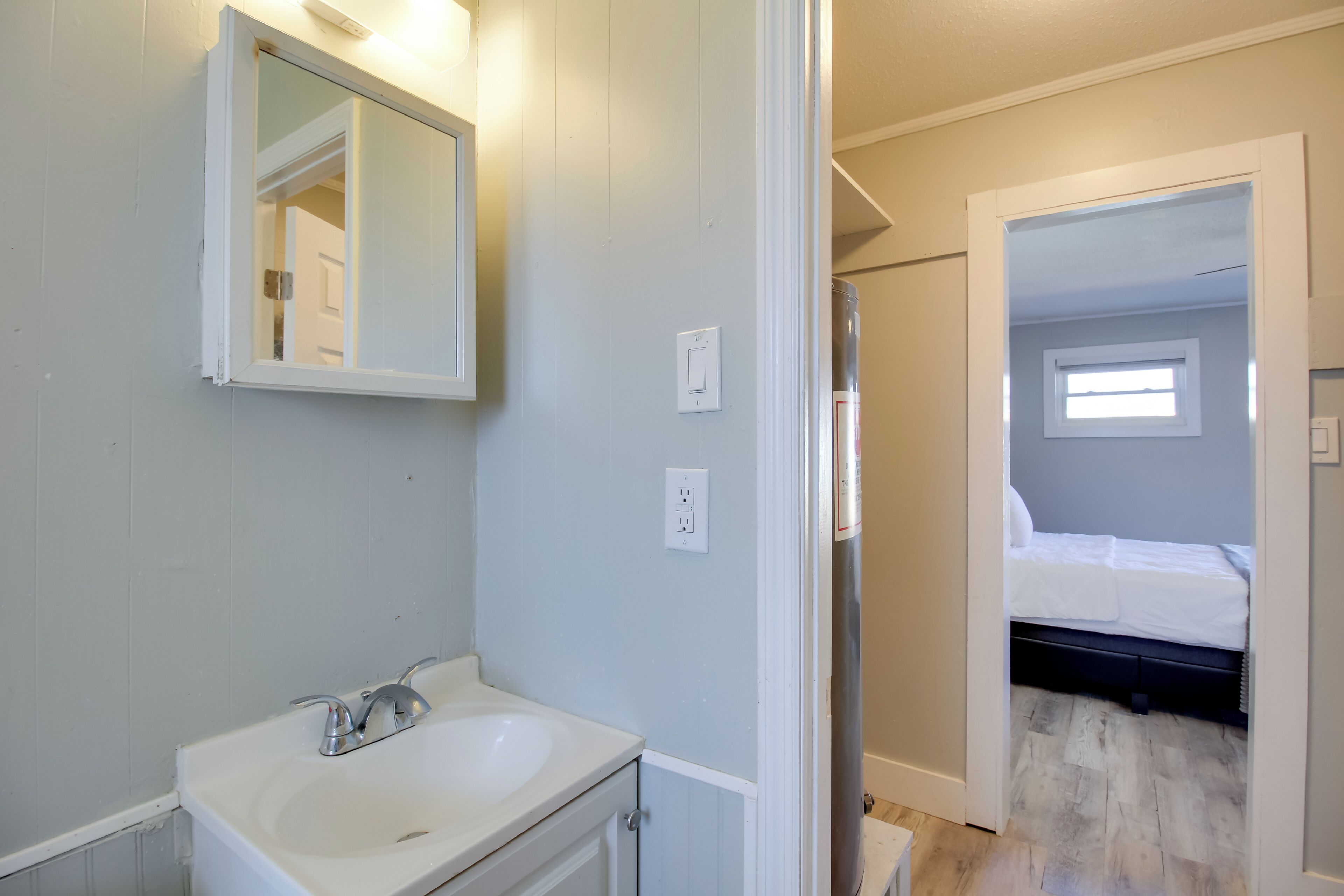 A bathroom with a white sink, mirror, and light switch. A bedroom is visible through an open door, featuring a bed and a window.
