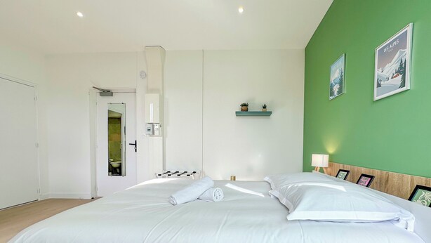 A bedroom with a large bed, a green accent wall, a small shelf with plants, and framed pictures. - #IH - Le Victoria 4 💟 (Grenoble)