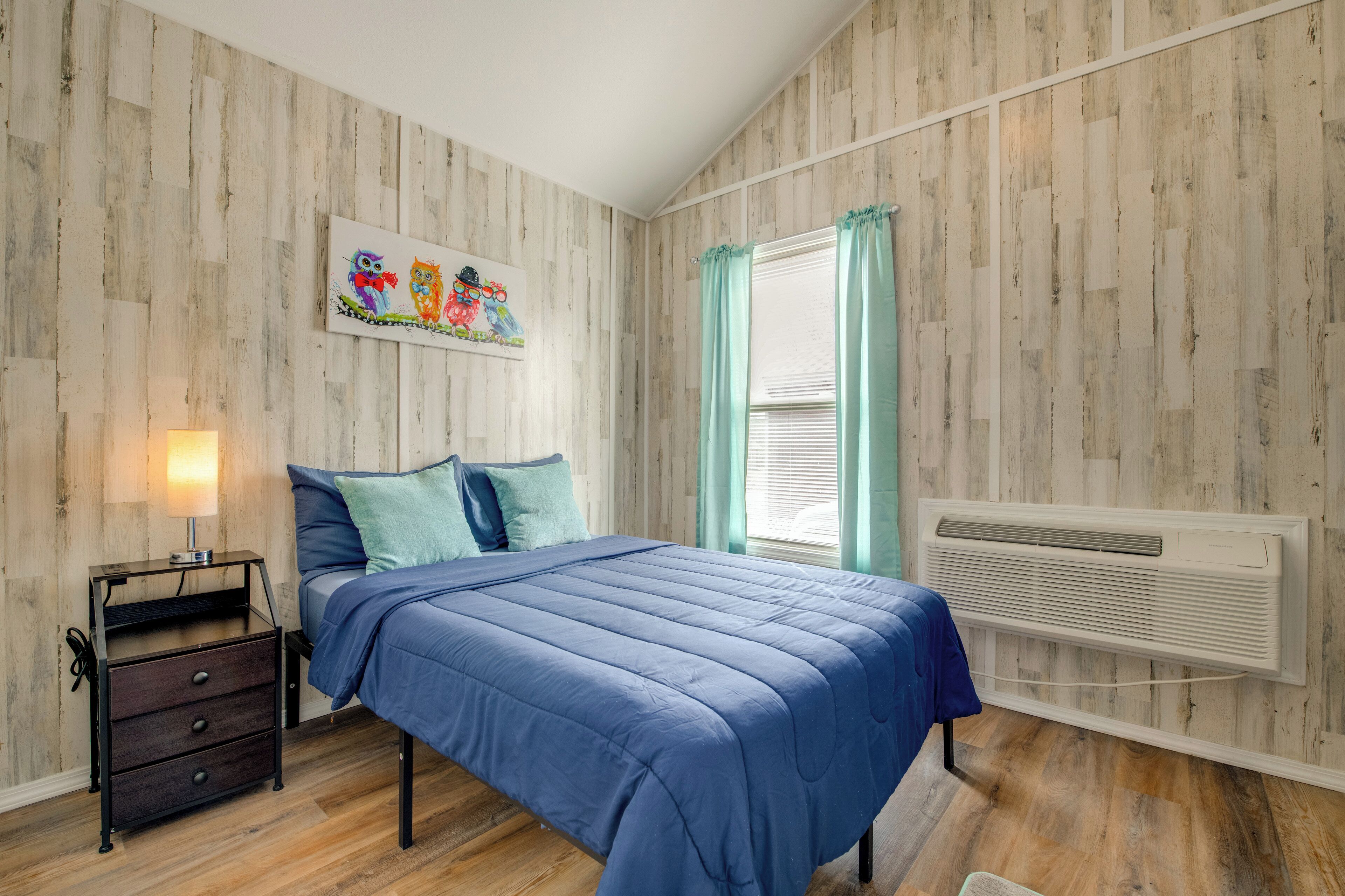 Ein Schlafzimmer mit Holzwandverkleidung, einem Bett mit blauer Bettwäsche, einem Nachttisch mit Schubladen, einem Fenster mit Vorhängen und einer wandmontierten Klimaanlage.