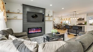 Uma sala de estar moderna com uma televisão de tela plana, sofás cinzas, uma mesa de centro e uma cozinha ao fundo.