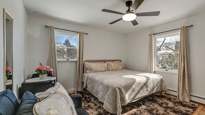 Quarto com cama, ventoinha no teto, janela com cortinas, mesinha com um vaso de flores e um sofá.