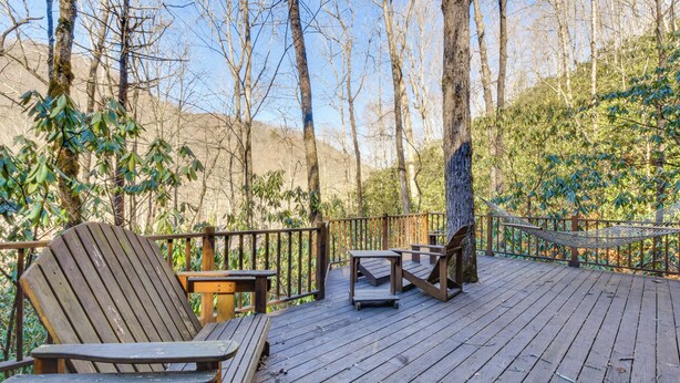 Una terraza de madera con dos sillas Adirondack y una pequeña mesa, rodeada de árboles y una hamaca.