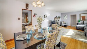 Un coin repas avec une table en bois dressée pour quatre personnes, entourée de chaises à motifs. Un lustre est suspendu au plafond, et une horloge est visible au mur.