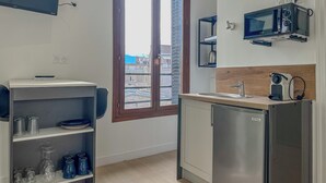 Une cuisine compacte équipée d’un four à micro-ondes encastré, d’un évier et d’un petit réfrigérateur. Une fenêtre offre une vue sur les bâtiments environnants.
