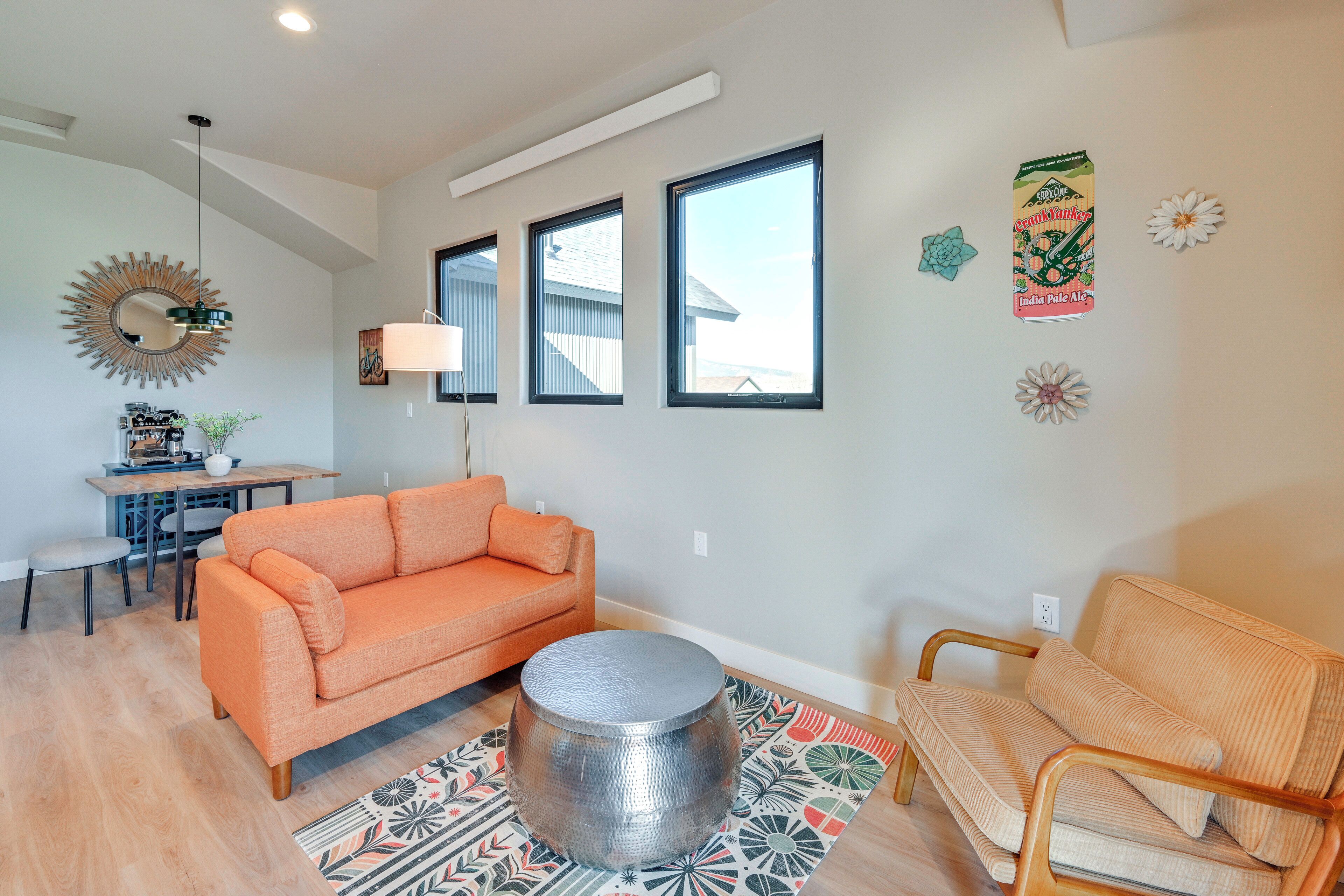 A living room with a sofa, a round coffee table, and a patterned rug. There are three windows and a mirror on the wall.