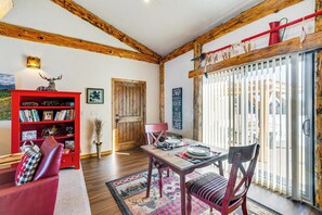 Een gezellige woonkamer met een rode boekenkast, een eettafel met stoelen, een open haard en een groot raam met gordijnen.