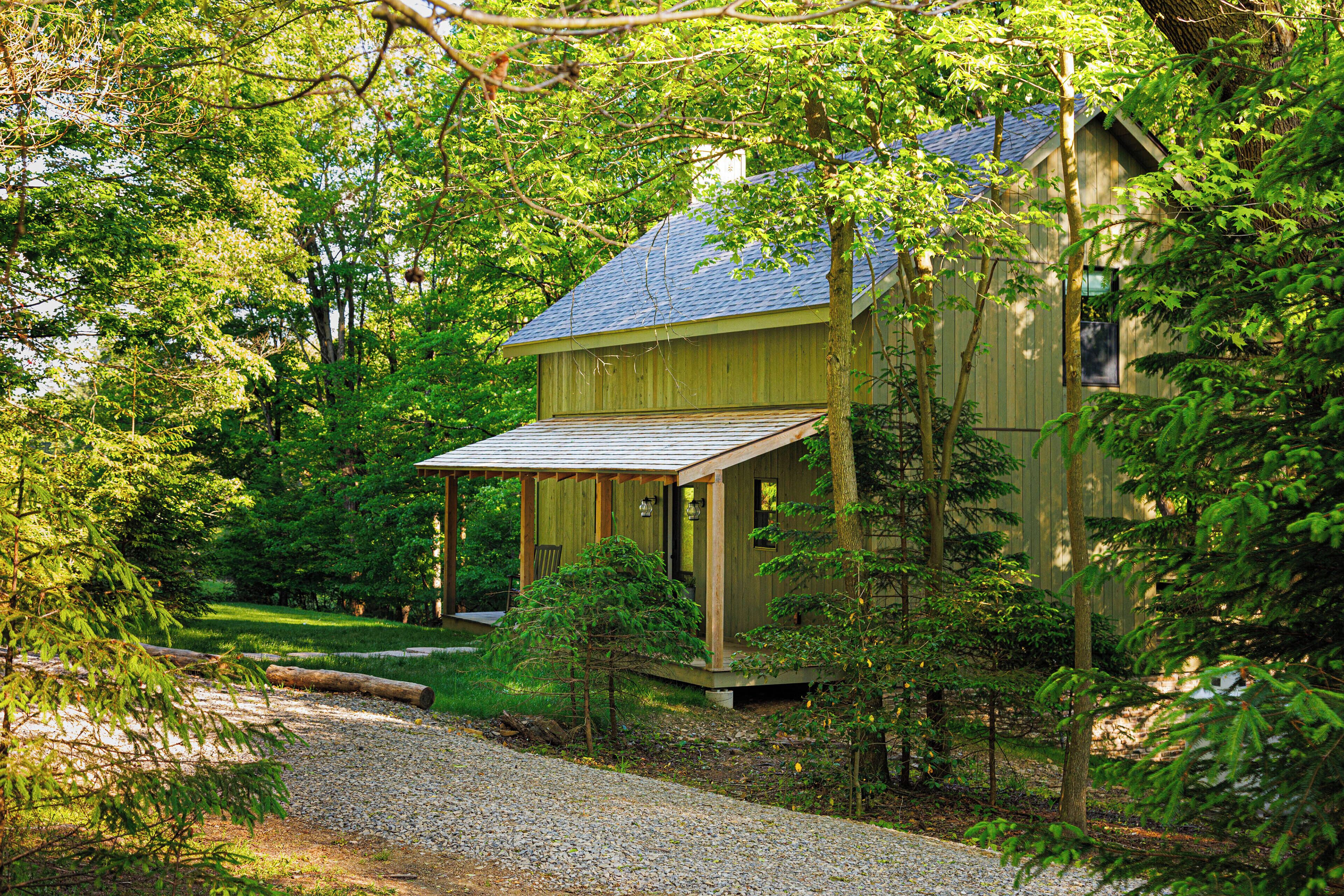 A wooden cabin with a porch, surrounded by dense trees and a gravel path.