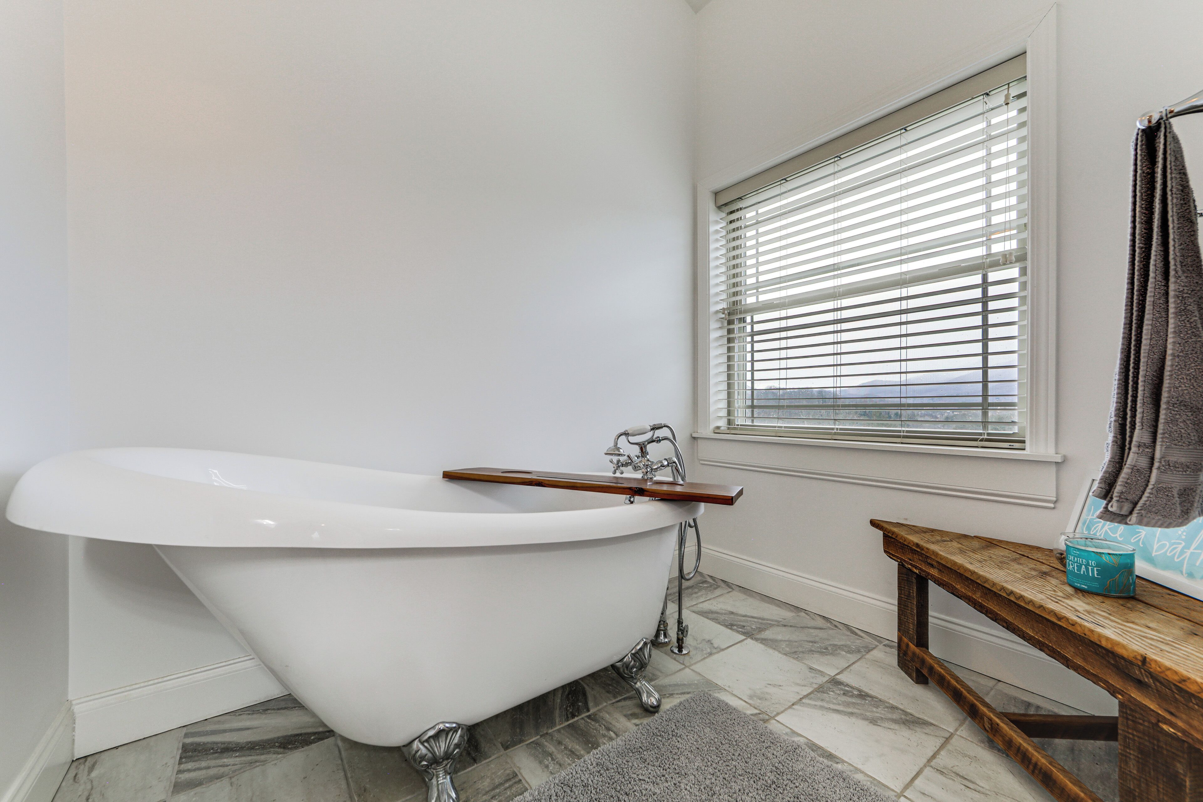 A bathroom with a freestanding bathtub, a wooden bench, and a window with blinds.