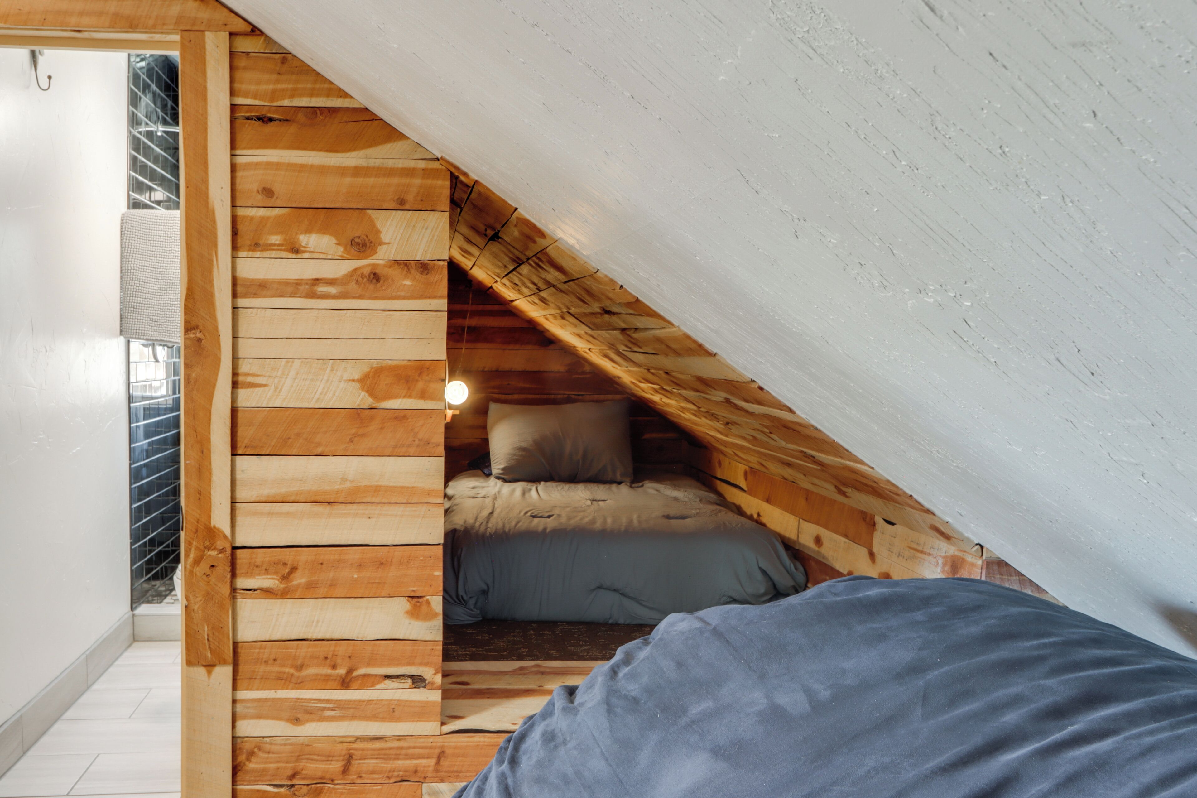 Un dormitorio compacto con techo inclinado, paneles de madera, cama con ropa de cama gris y una ventana pequeña.