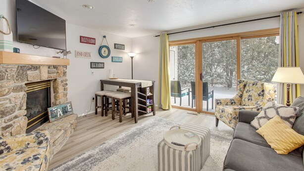 Una sala de estar acogedora con chimenea de piedra, un televisor de pantalla plana, una mesa de comedor con sillas, un sofá con cojines y un ventanal con cortinas.