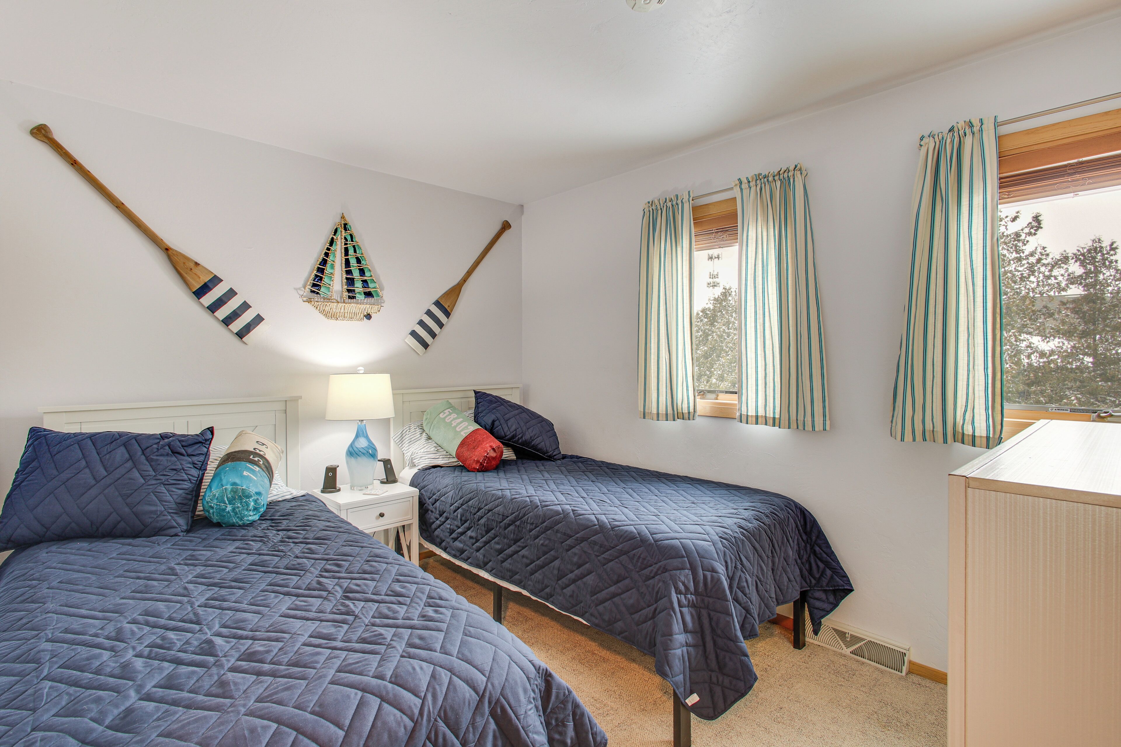 A bedroom with two beds, a nightstand, a lamp, and two windows with curtains.