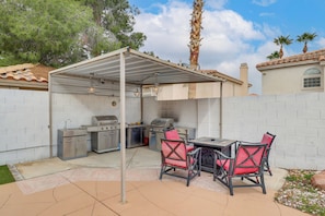 Un patio exterior con zona de parrilla cubierta, una mesa con sillas y un muro de ladrillo.